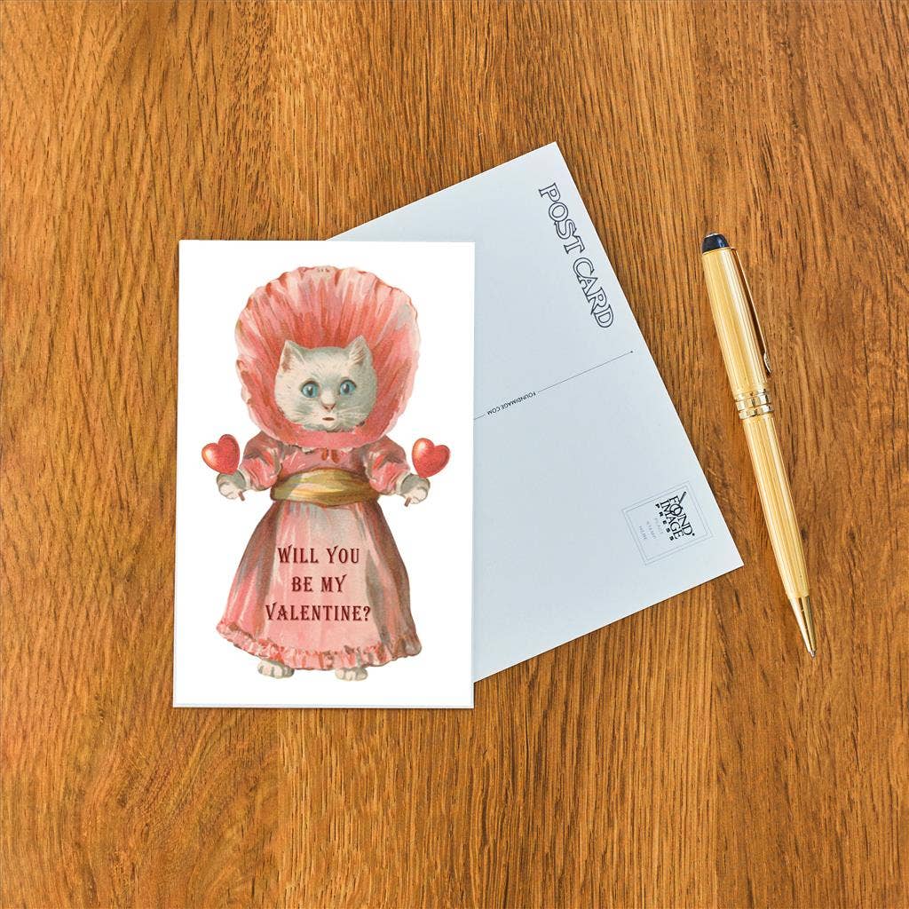 Valentine's Day card with a cat in a pink dress and bonnet on a wooden surface with a pen.