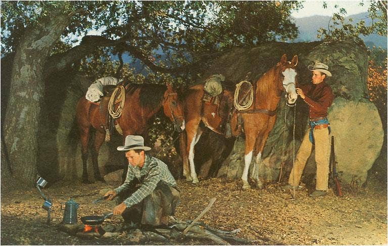 Two cowboys tending to their horses under a tree, with one cooking on a fire.