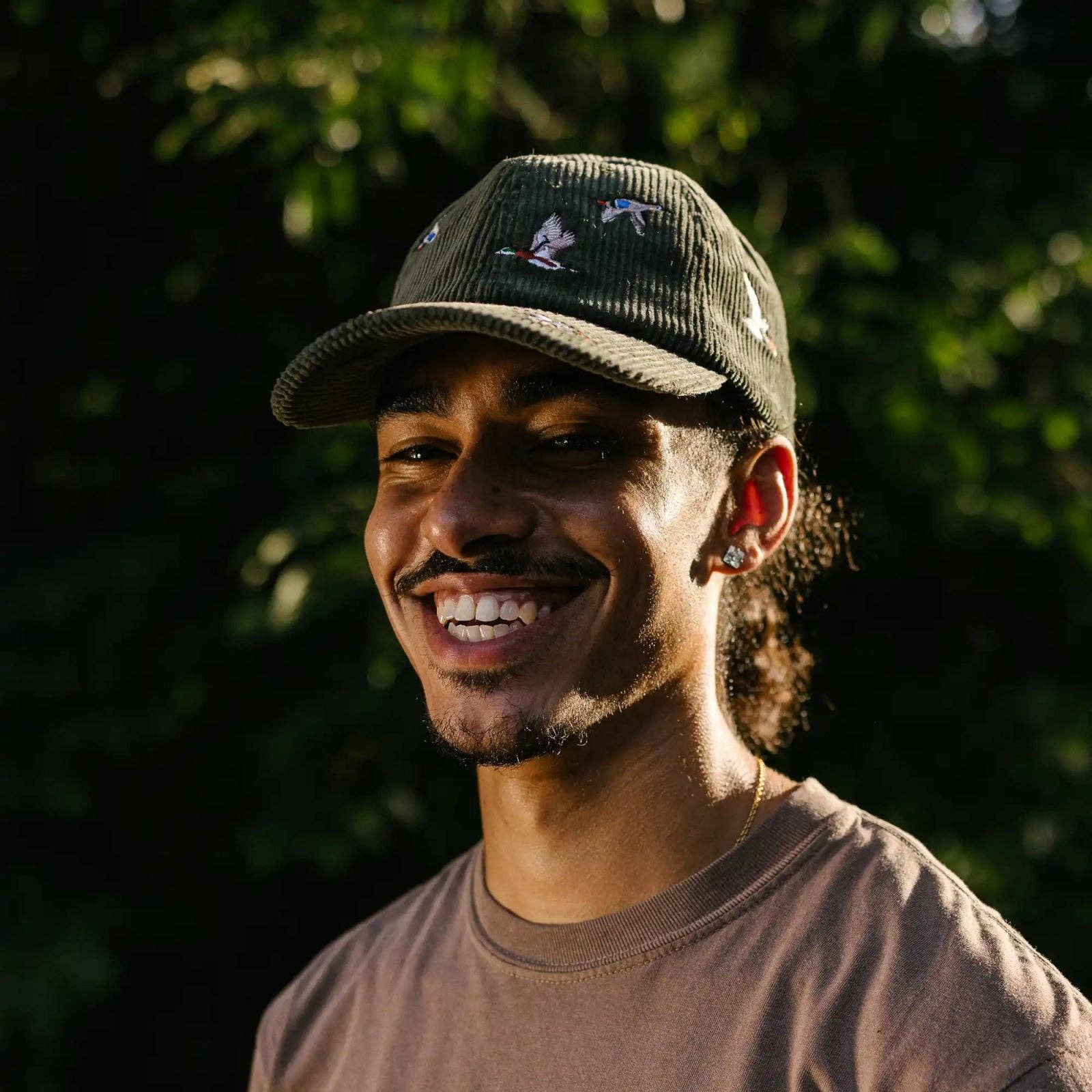 Man wearing a cap and smiling outdoors with greenery in the background