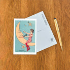 Vintage-style postcard with a woman on the moon, pen, and envelope on a wooden surface