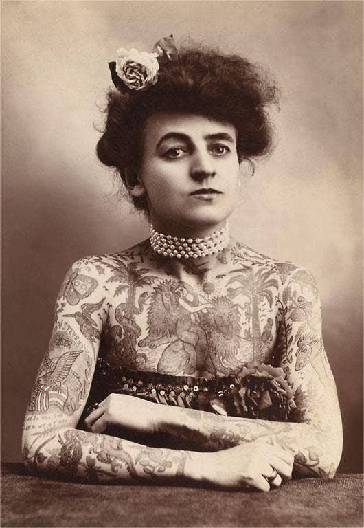 Vintage photograph of a woman with tattoos on her arms, wearing a pearl necklace and a decorative headpiece.