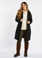 Woman wearing a long black coat, white sweater, green pants, and brown knee-high boots on a white background