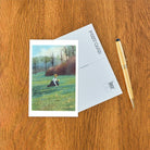 Postcard with a scenic illustration of a person in a field, accompanied by an envelope and a pen on a wooden surface.