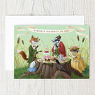 Birthday card with illustrated animals around a tree stump on a light wooden background