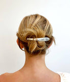 Back view of a person with blonde hair styled in an updo, wearing a silver hair clip against a white background.