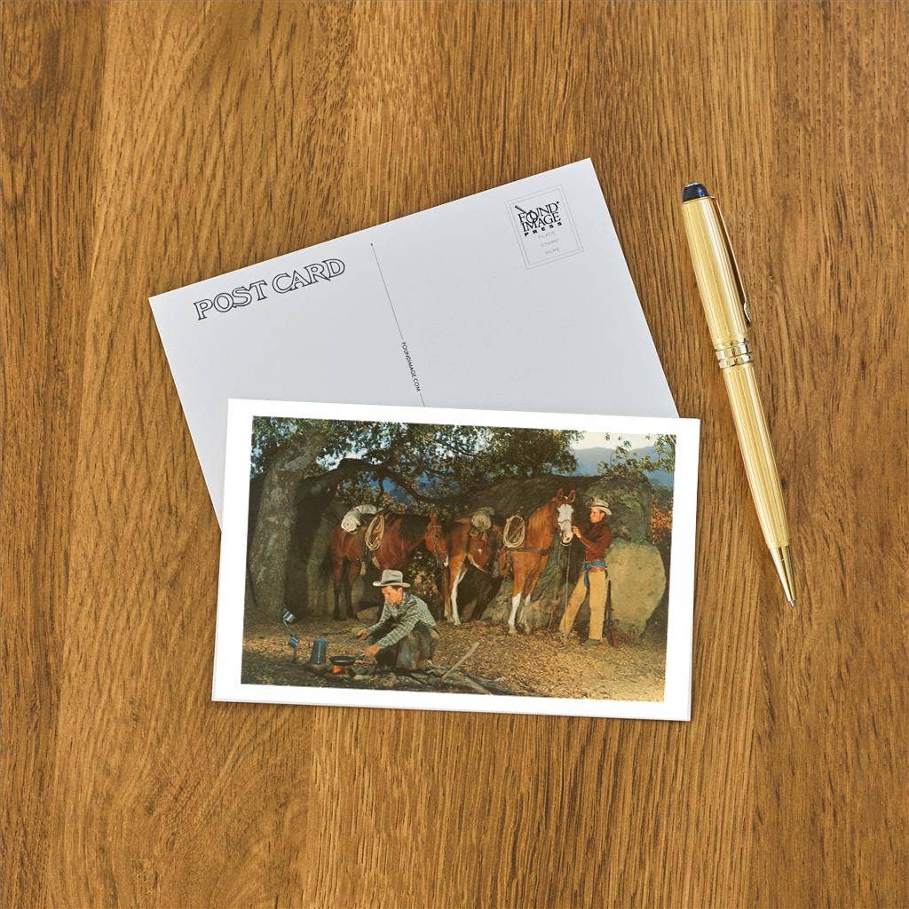 Postcard with a painting of cowboys and horses on a wooden surface with a pen.